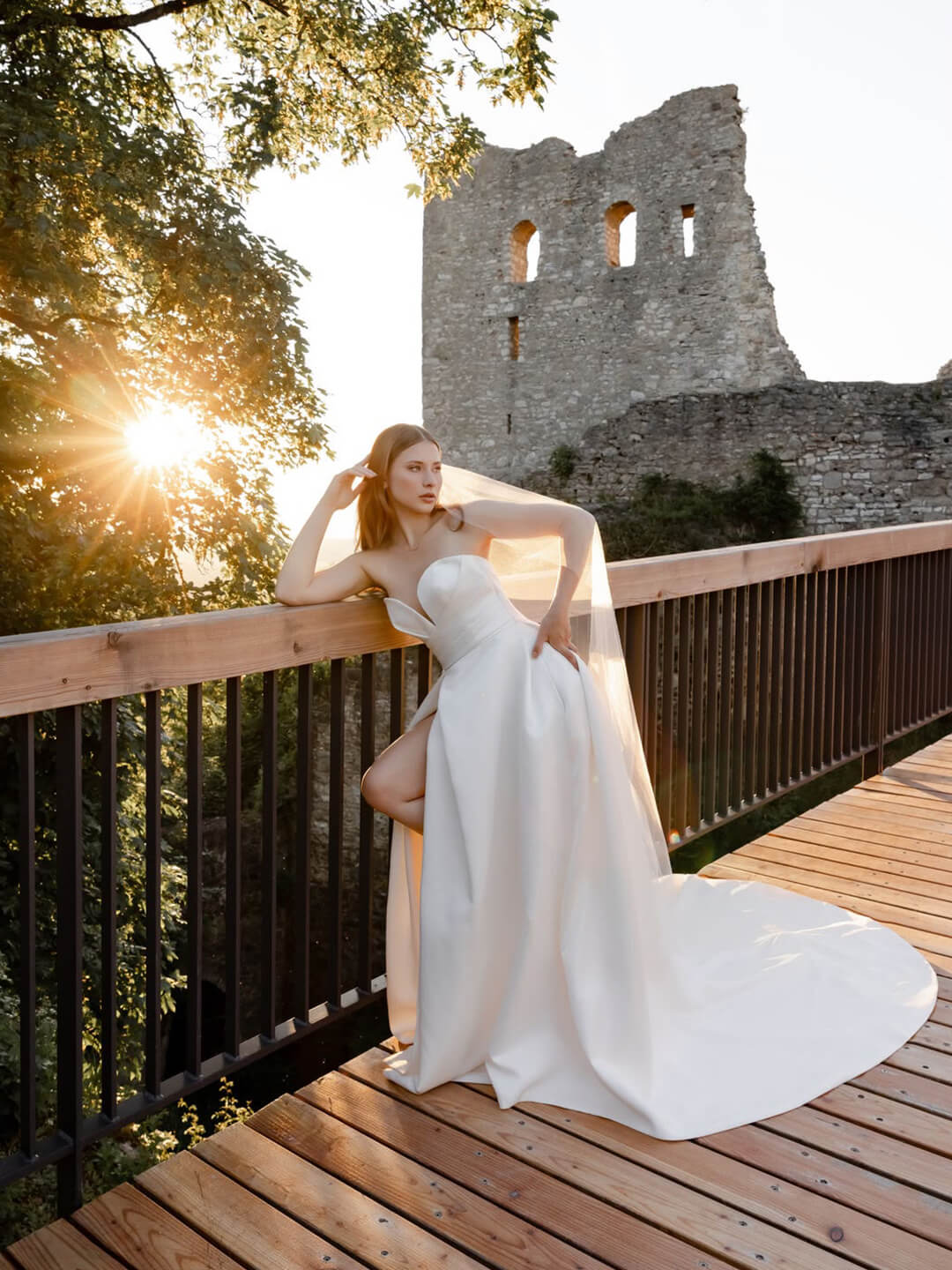 Braut in einem weißen Brautkleid mit herzförmigem Ausschnitt lehnt an einem Geländer einer Holzbrücke. Das Kleid hat einen hohen Beinschlitz und eine lange Schleppe. Im Hintergrund sind eine Burgruine und die tief stehende Sonne zu sehen.