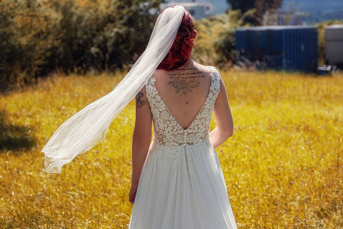 Rückenansicht einer Braut in einem weißen Brautkleid mit tiefem Rückenausschnitt und langem Schleier auf einer Wiese. Der Schleier weht im Wind.