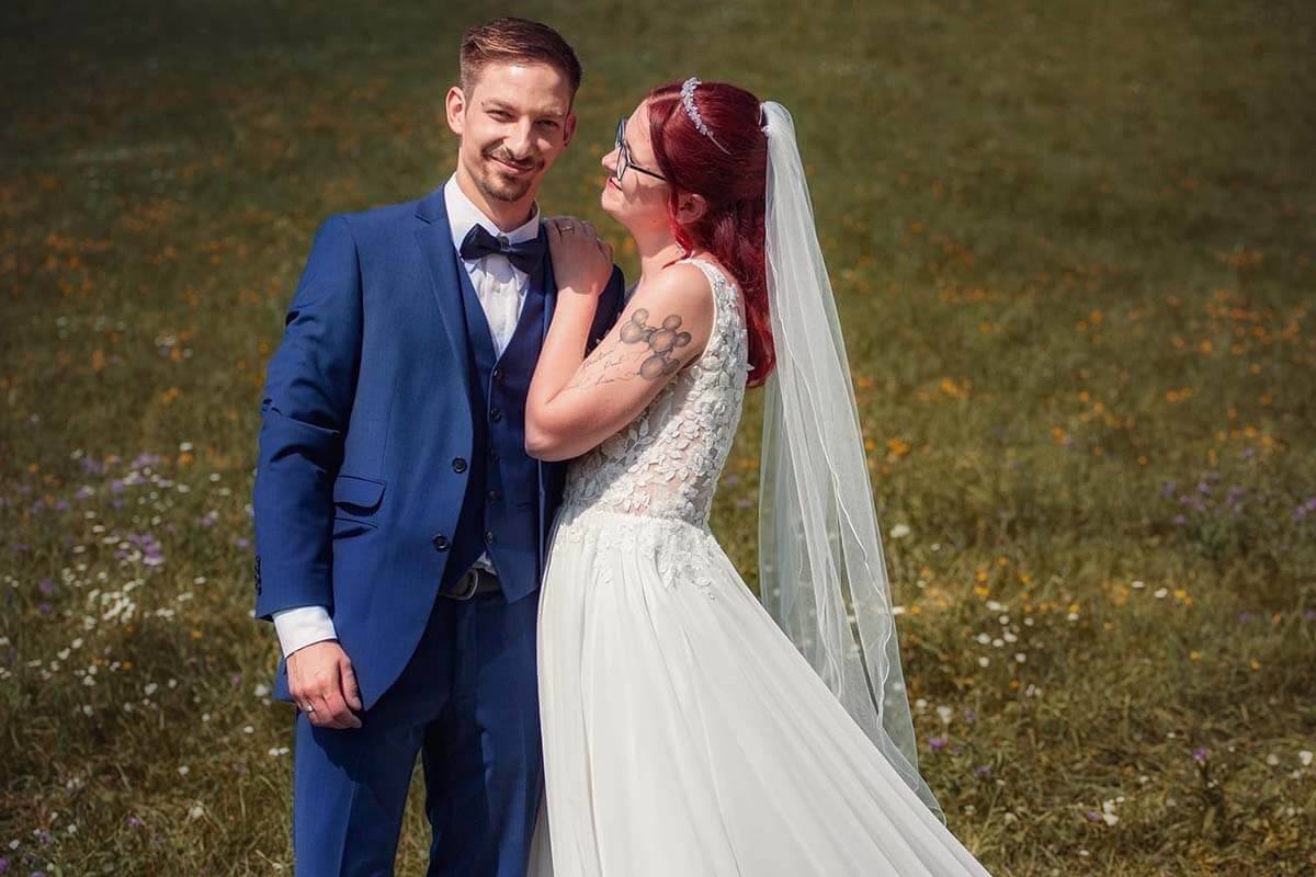 Bräutigam im blauen Anzug steht lächelnd neben seiner Braut mit roten Haaren, Brille und weißem Brautkleid mit langer Schleppe auf einer Blumenwiese